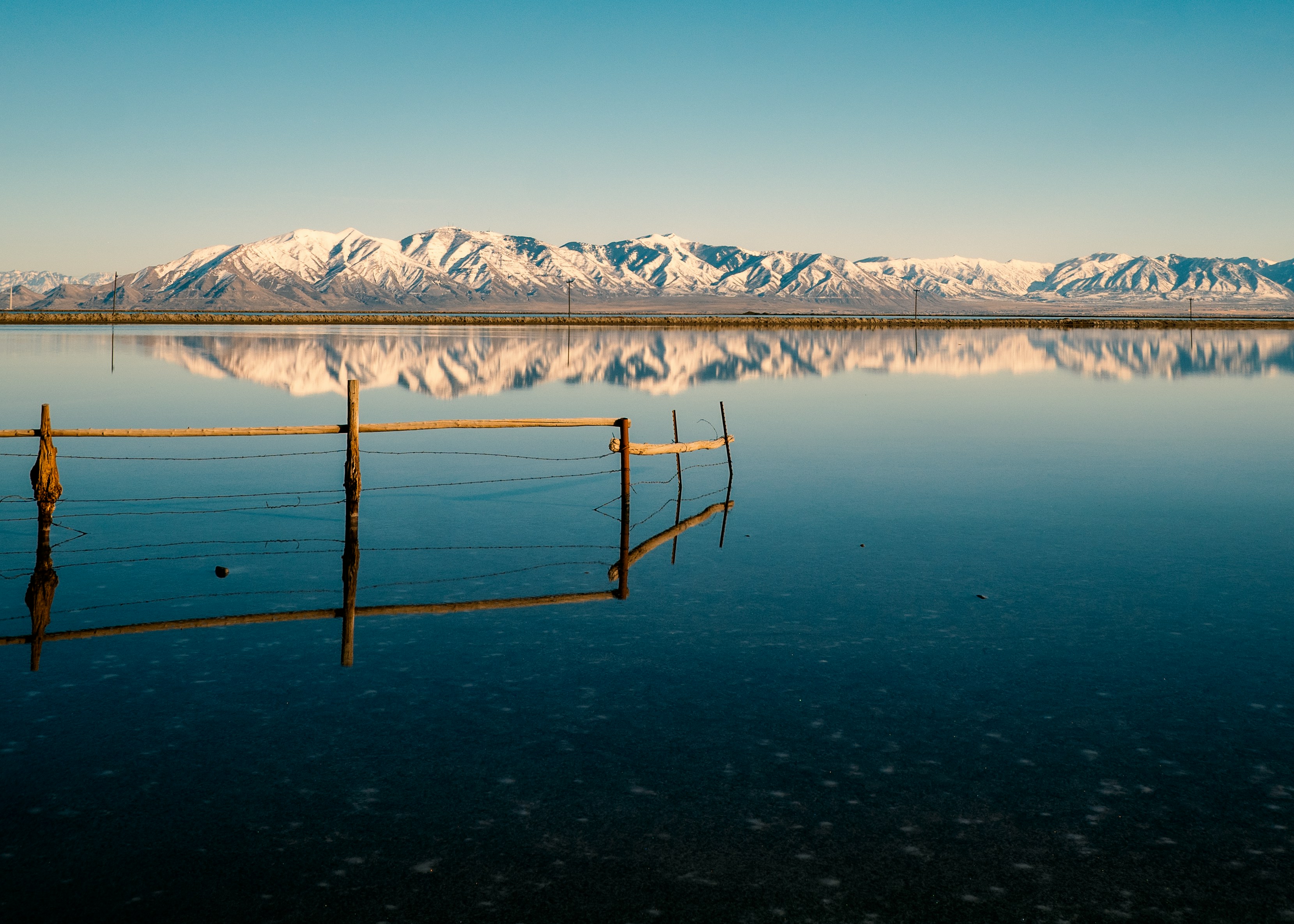 A submerged fence line mirrored in the still surface of the lake, snow-capped Wasatch behind